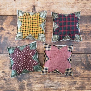 May include: Four square decorative pillows with star-shaped designs. The pillows feature various patterns, including yellow plaid, red and green plaid, burgundy with white stars, and red gingham. Each pillow has a patterned border.