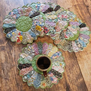 May include: A collection of handmade, quilted flower-shaped coasters in various colors and patterns. One coaster holds a cup of coffee. The coasters have a green center and scalloped edges. The background is a wooden surface.