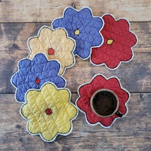 May include: A collection of quilted flower-shaped coasters in various colors, including red, yellow, and blue. Each coaster has a contrasting center and a white border with black stitching. One coaster holds a cup of coffee.