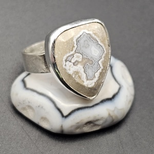 May include: A silver ring with a large, shield-shaped stone. The stone features a beige and gray patterned design, set in a silver bezel. The ring is displayed on a white and gray patterned stone.