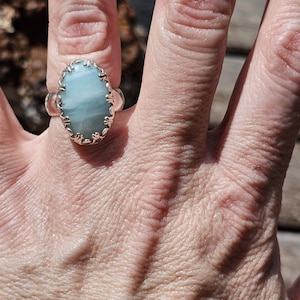 May include: A silver ring featuring an oval-shaped, light blue stone with subtle white horizontal banding. The stone is held in place by a decorative, scalloped metal bezel setting that encircles the entire perimeter of the gemstone.