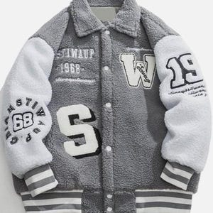 Puede incluir: Una chaqueta de estilo universitario gris y blanca hecha de material de forro polar texturizado. La chaqueta cuenta con un frente abotonado, un cuello y mangas blancas. Muestra el texto STIWAUP 1968, la letra W, la letra S y el número 19.