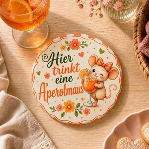 Könnte beinhalten: Ein runder Keramikuntersetzer mit einer gezeichneten Maus mit gepunkteter Schleife, die einen orangefarbenen Cocktail hält. Der Text lautet Hier trinkt eine Aperolmaus. Das Design zeigt orangefarbene und gelbe Blumen, grüne Blätter und kleine Herzen.
