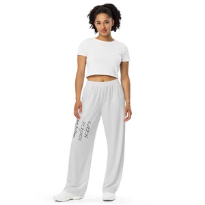 May include: A woman wearing a white crop top and white sweatpants. The sweatpants have the text "CATCH FLIGHTS not feelings" printed in black on the left leg. The woman is wearing white sneakers.