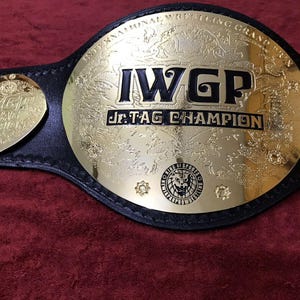 Könnte beinhalten: Ein gold-schwarzer Wrestling-Meisterschaftsgürtel. Der Gürtel hat eine große goldene Mittelplatte mit dem Text "IWGP Jr. Tag Champion". Der Gürtel hat zwei kleinere goldene Platten und einen schwarzen Lederriemen.