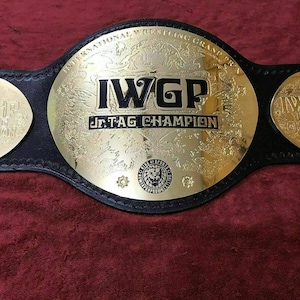 Könnte beinhalten: Ein schwarzer Leder-Meisterschaftsgürtel mit einer großen, goldfarbenen Mittelplatte, die den Text IWGP Jr. TAG CHAMPION und ein kleines Löwenlogo trägt. Zwei kleinere, goldfarbene runde Platten sind an den Seiten mit gleichem Text befestigt.