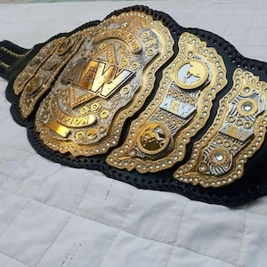 May include: A championship wrestling belt featuring a black leather strap with intricate gold and silver metal plates. The central plate displays the text AEW World Champion with decorative engravings and embedded clear stones throughout the design.