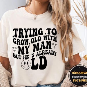 May include: Cream-colored t-shirt with the text "TRYING TO GROW OLD WITH MY MAN BUT HE'S ALREADY OLD" in black, retro-style font. The design includes hearts and a smiley face. The shirt is a casual wear item.