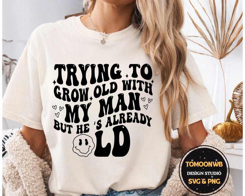 May include: Cream-colored t-shirt with the text "TRYING TO GROW OLD WITH MY MAN BUT HE'S ALREADY OLD" in black, retro-style font. The design includes hearts and a smiley face. The shirt is a casual wear item.