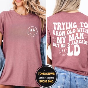 May include: A mauve-colored t-shirt with a white smiley face graphic on the front and the text "TRYING TO GROW OLD WITH MY MAN BUT HE'S ALREADY OLD" on the back. The shirt is a casual style.