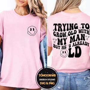 May include: A light pink t-shirt with a black smiley face graphic on the front and the text "TRYING TO GROW OLD WITH MY MAN BUT HE'S ALREADY OLD" on the back. The shirt is a casual style.