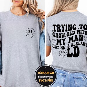 May include: A light gray t-shirt with a black smiley face graphic on the front and the text "TRYING TO GROW OLD WITH MY MAN BUT HE'S ALREADY OLD" on the back. The shirt is a casual style.