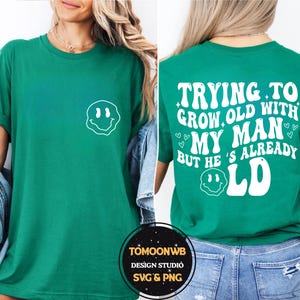 May include: Green t-shirt with a white smiley face graphic on the front. The back of the shirt features the text "TRYING TO GROW OLD WITH MY MAN BUT HE'S ALREADY OLD" in a retro font. A casual, unisex tee.
