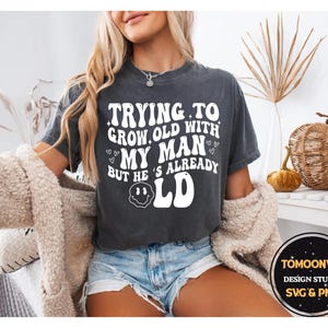 May include: Dark gray t-shirt with the text "TRYING TO GROW OLD WITH MY MAN BUT HE'S ALREADY OLD" in a retro font. The shirt also features a smiley face and heart graphics. The model is wearing blue jean shorts.