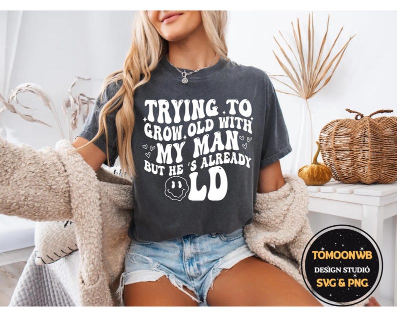 May include: Dark gray t-shirt with the text "TRYING TO GROW OLD WITH MY MAN BUT HE'S ALREADY OLD" in a retro font. The shirt also features a smiley face and heart graphics. The model is wearing blue jean shorts.