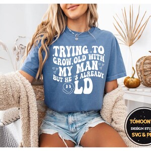 May include: A blue t-shirt with the text "Trying to grow old with my man but he's already old" in white. The shirt has a smiley face graphic and is paired with denim shorts. A cozy beige sweater is draped over the shoulders.