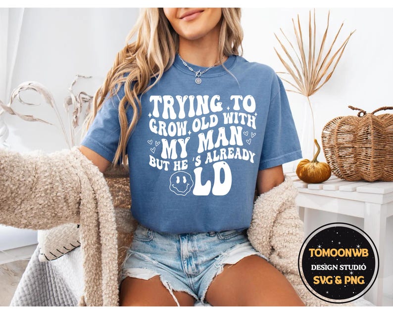 May include: A blue t-shirt with the text "Trying to grow old with my man but he's already old" in white. The shirt has a smiley face graphic and is paired with denim shorts. A cozy beige sweater is draped over the shoulders.