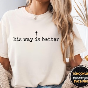 May include: A cream-coloured t-shirt featuring the text "his way is better" in black, with a cross symbol above the word "is". The shirt is a casual style, suitable for everyday wear.