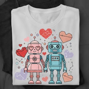 Może przedstawiać: Biała koszulka z kreskówkową ilustracją dwóch robotów trzymających się za ręce, otoczonych kształtami serc i konstelacjami. Roboty są różowe i niebieskie, z oczami w kształcie serca i retro designem. Tematem jest miłość i romans.