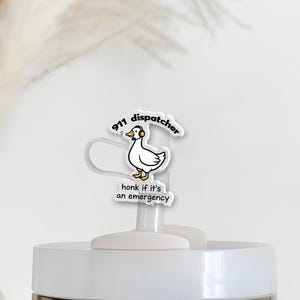 Puede incluir: Un vaso blanco con tapa blanca y plateada, coronado por un dije de plástico transparente. El dije presenta un pato de dibujos animados con auriculares, con el texto "911 dispatcher" y "honk if it's an emergency".