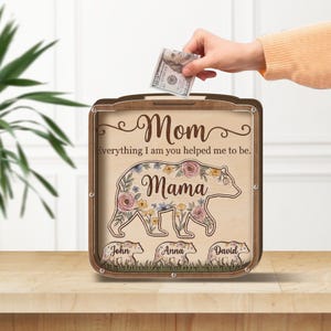 May include: A wooden money box with the words "Mom" and "Mama" in script. The box features a bear design with floral accents and the names "John", "Anna", and "David". A hand is inserting a $100 bill into the slot.
