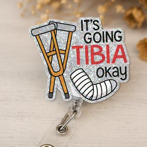 May include: A badge reel with a glittery silver background. The design includes yellow crutches, a bandaged leg, and the text "It's Going Tibia Okay" in black and red. The reel has a clear plastic strap and a metal clip.