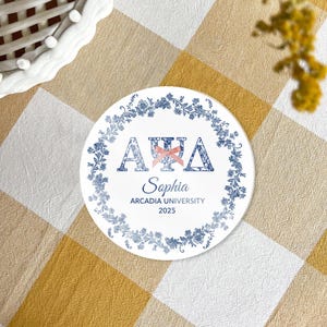 May include: A white round coaster with blue floral accents and text. The text includes the Greek letters Alpha, Chi, and Delta, the name "Sophia", "Arcadia University", and the year "2025". A pink bow is between the Greek letters.