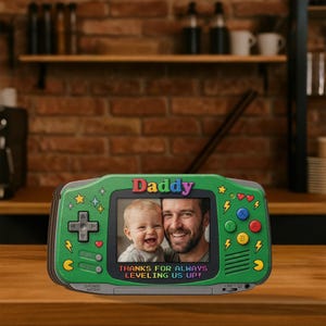 May include: A green picture frame shaped like a retro handheld game console. The frame has a photo of a man and a baby. The word "Daddy" is at the top, and the text "THANKS FOR ALWAYS LEVELING US UP!" is at the bottom.