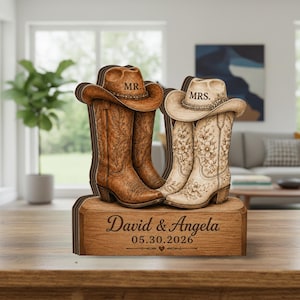 May include: Wooden wedding decoration featuring two pairs of cowboy boots and hats. One pair is brown with "MR." on the hat, the other is cream-colored with "MRS." on the hat. The base is engraved with "David & Angela" and the wedding date.