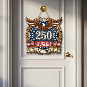 K&ouml;nnte beinhalten: Ein dekoratives Holzschild f&uuml;r die Wand mit einem Wei&szlig;kopfseeadler mit ausgebreiteten Fl&uuml;geln &uuml;ber der Zahl 250, dem Text UNITED STATES OF AMERICA und den Jahren 1776-2026. Das Design umfasst einen blauen Hintergrund mit wei&szlig;en Sternen, goldenen Lorbeerkr&auml;nzen und Flaggen.