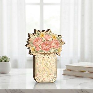 May include: Wooden mason jar decoration with a floral arrangement of pink and yellow flowers. The jar features the letter "S" and a floral pattern. The decoration is made of wood and has a brown border.