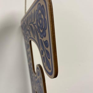 K&ouml;nnte beinhalten: Ein flacher, buchstabenf&ouml;rmiger Wandbehang aus Holz mit hellbrauner Oberfl&auml;che und einem blauen Blumenmuster an den R&auml;ndern. Das Objekt hat ein glattes, geschichtetes Holzprofil an der Seite und ist an einem d&uuml;nnen, wei&szlig;en geflochtenen Seil aufgeh&auml;ngt.