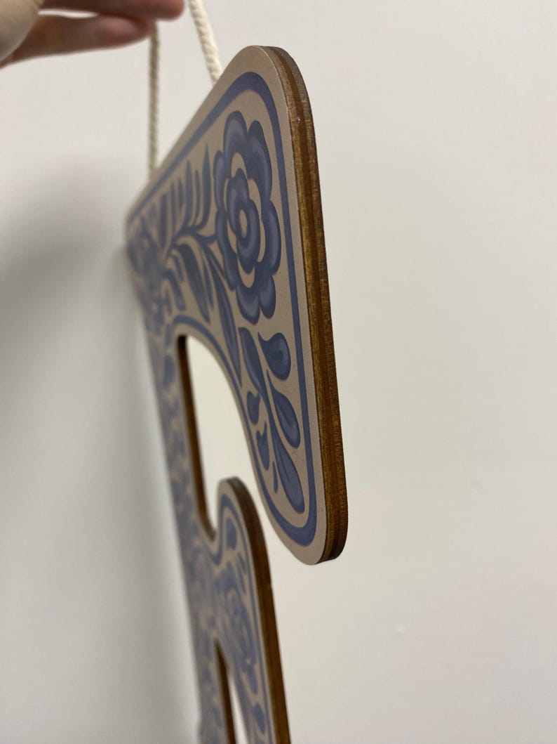 K&ouml;nnte beinhalten: Ein flacher, buchstabenf&ouml;rmiger Wandbehang aus Holz mit hellbrauner Oberfl&auml;che und einem blauen Blumenmuster an den R&auml;ndern. Das Objekt hat ein glattes, geschichtetes Holzprofil an der Seite und ist an einem d&uuml;nnen, wei&szlig;en geflochtenen Seil aufgeh&auml;ngt.