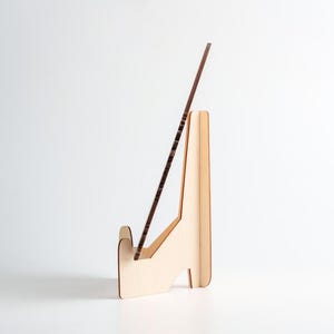 K&ouml;nnte beinhalten: Ein heller Holzst&auml;nder mit einer vertikalen R&uuml;ckenst&uuml;tze und einer vorderen Ablage, der einen einzelnen d&uuml;nnen, dunkelbraunen, rechteckigen Holzstab in einem Winkel h&auml;lt. Der St&auml;nder besteht aus flachen, lasergeschnittenen Holzteilen.