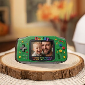 May include: A green photo frame shaped like a retro video game console. The frame has a photo of a man and a baby. The word "Daddy" is at the top, and the text "THANKS FOR ALWAYS LEVELING US UP!" is at the bottom.