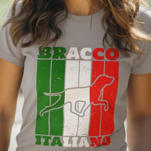 Può includere: T-shirt grigio chiaro con la bandiera italiana e la silhouette di un cane Bracco Italiano. Le parole "BRACCO" e "ITALIANO" sono stampate in verde e rosso sulla maglietta.