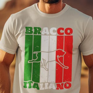 Può includere: T-shirt grigio chiaro con la scritta "BRACCO ITALIANO" in verde e rosso, e un contorno bianco di un cane. Il design utilizza i colori della bandiera italiana. La maglietta è a girocollo.