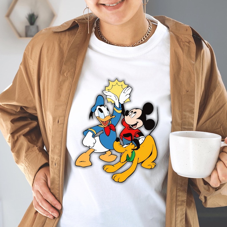Peut inclure: T-shirt blanc avec un graphisme color&eacute; des personnages Disney Donald Duck, Mickey Mouse et Pluto. Donald Duck porte un costume de marin bleu et Mickey Mouse une chemise rouge. Pluto est jaune. Le graphisme comprend un soleil jaune.