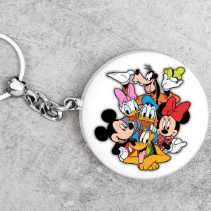 Peut inclure: Un porte-cl&eacute;s rond blanc avec une illustration color&eacute;e des personnages Disney Mickey Mouse, Minnie Mouse, Donald Duck, Daisy Duck, Dingo et Pluto. Le porte-cl&eacute;s a une cha&icirc;ne et un anneau en m&eacute;tal argent&eacute;.