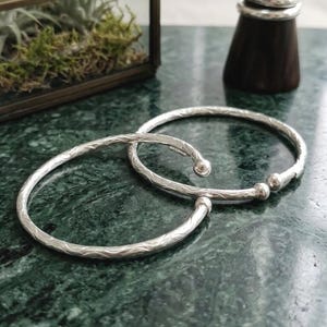 May include: Two silver bangles with a textured, wave-like pattern. One bangle has a rounded ball on each end, while the other has a single ball. The bangles are displayed on a green marble surface, with a small terrarium and wooden ring holder in the background.