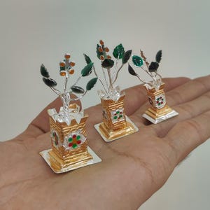 Può includere: Tre sculture decorative di piante in miniatura in argento e oro. Ognuna ha una base quadrata dorata e un vaso dorato con disegni floreali rossi, verdi e bianchi. Le piante hanno steli argentati con foglie verdi e boccioli floreali arancioni e bianchi.