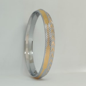 May include: A silver-colored bangle with a gold-colored, textured band. The gold band features a repeating chevron pattern. The bangle is a simple, elegant piece of jewelry.
