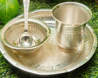May include: A silver puja thali set, comprising a small bowl with a spoon, a cup, and a plate. The plate features a honeycomb pattern and a decorative edge. The set is presented on green grass.