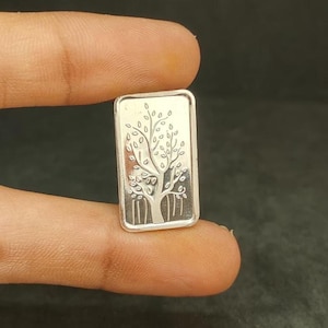 May include: A small, rectangular silver bar with a detailed tree design. The tree has a trunk and branches with leaves. The bar is held between fingers, showcasing the intricate engraving.