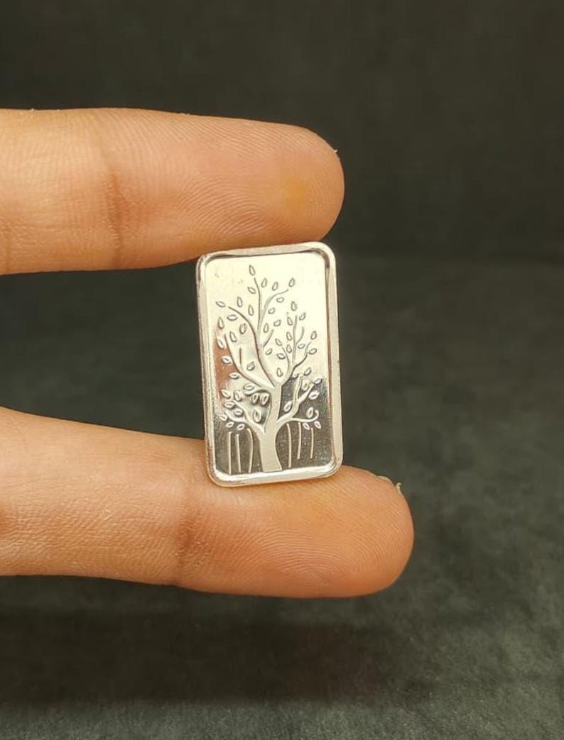 May include: A small, rectangular silver bar with a detailed tree design. The tree has a trunk and branches with leaves. The bar is held between fingers, showcasing the intricate engraving.