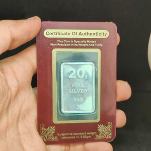 May include: A silver bar, marked '20 FINE SILVER 999', is encased in a certificate of authenticity. The certificate, in a maroon frame, states the coin is specially minted with precision. The text 'subject to standard weight tolerance +/- 0.02gm' is also visible.