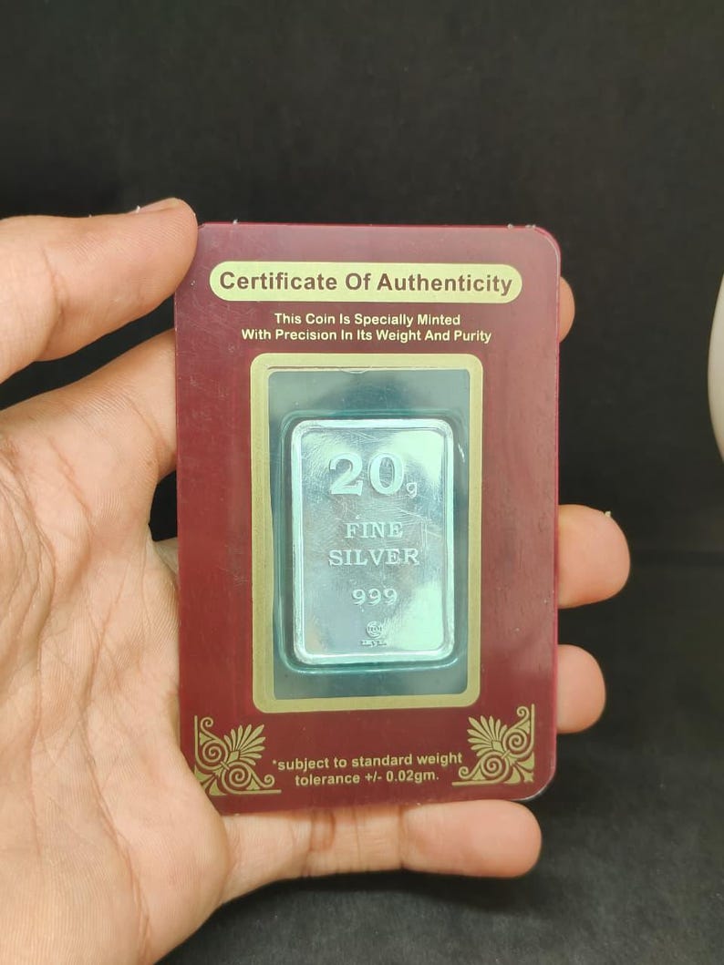 May include: A silver bar, marked '20 FINE SILVER 999', is encased in a certificate of authenticity. The certificate, in a maroon frame, states the coin is specially minted with precision. The text 'subject to standard weight tolerance +/- 0.02gm' is also visible.
