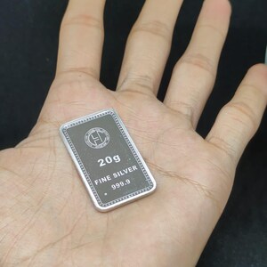 以下が含まれることがあります： 手のひらの上に置かれた、"20g FINE SILVER 999.9"と刻印された長方形の銀のバー。バーは、白い文字と銀色の縁取りのある濃い灰色の中心部を持っています。背景は暗い色です。