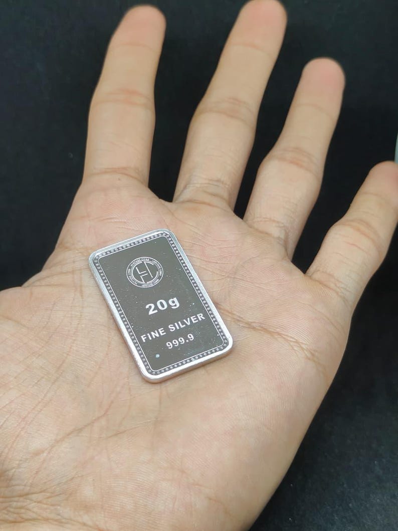 以下が含まれることがあります： 手のひらの上に置かれた、"20g FINE SILVER 999.9"と刻印された長方形の銀のバー。バーは、白い文字と銀色の縁取りのある濃い灰色の中心部を持っています。背景は暗い色です。