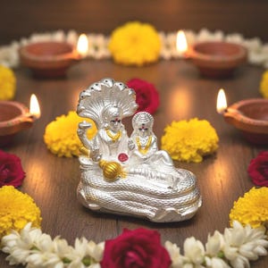 May include: A silver statue of two deities, adorned with gold necklaces, rests on a serpent base. The statue is surrounded by yellow and red flowers, white jasmine garlands, and lit clay lamps, creating a festive and spiritual ambiance.